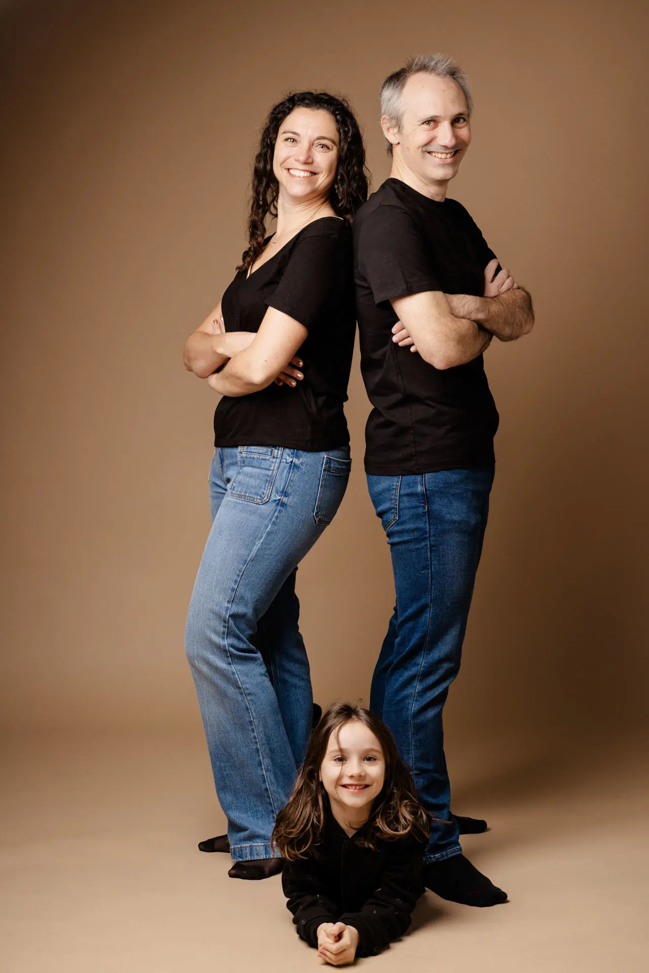 Séance photo famille en studio à Grenoble — parents dos à dos bras croisés, petite fille allongée au sol, tenues coordonnées noir et jeans, fond caramel — Utopikphoto