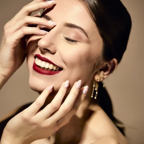Shooting beauté en studio à Grenoble — portrait serré femme brune mains sur le visage, rouge à lèvres bordeaux mat, ongles nacrés nail art et boucles dorées — photographe mode Utopikphoto